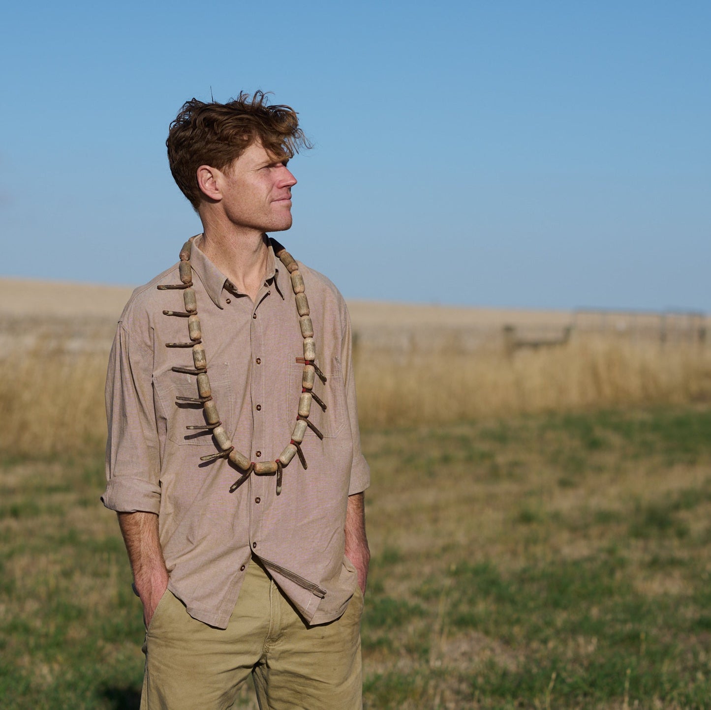 Man standing in a field wearing a beige shirt with decorative elements and green pants.