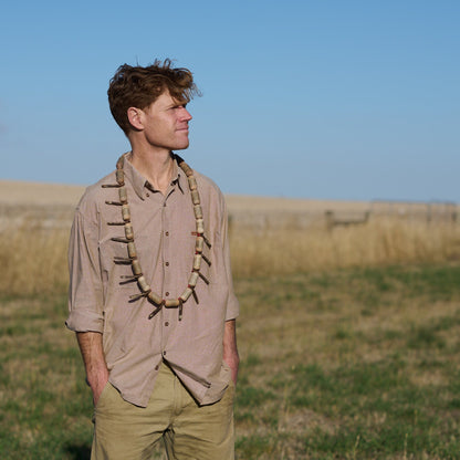 Man standing in a field wearing a beige shirt with decorative elements and green pants.