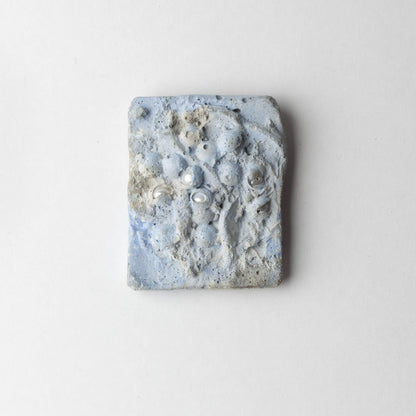 A square handmade ceramic brooch with a textured, organic surface featuring light blue, gray, and white marbled patterns, photographed against a clean white background.