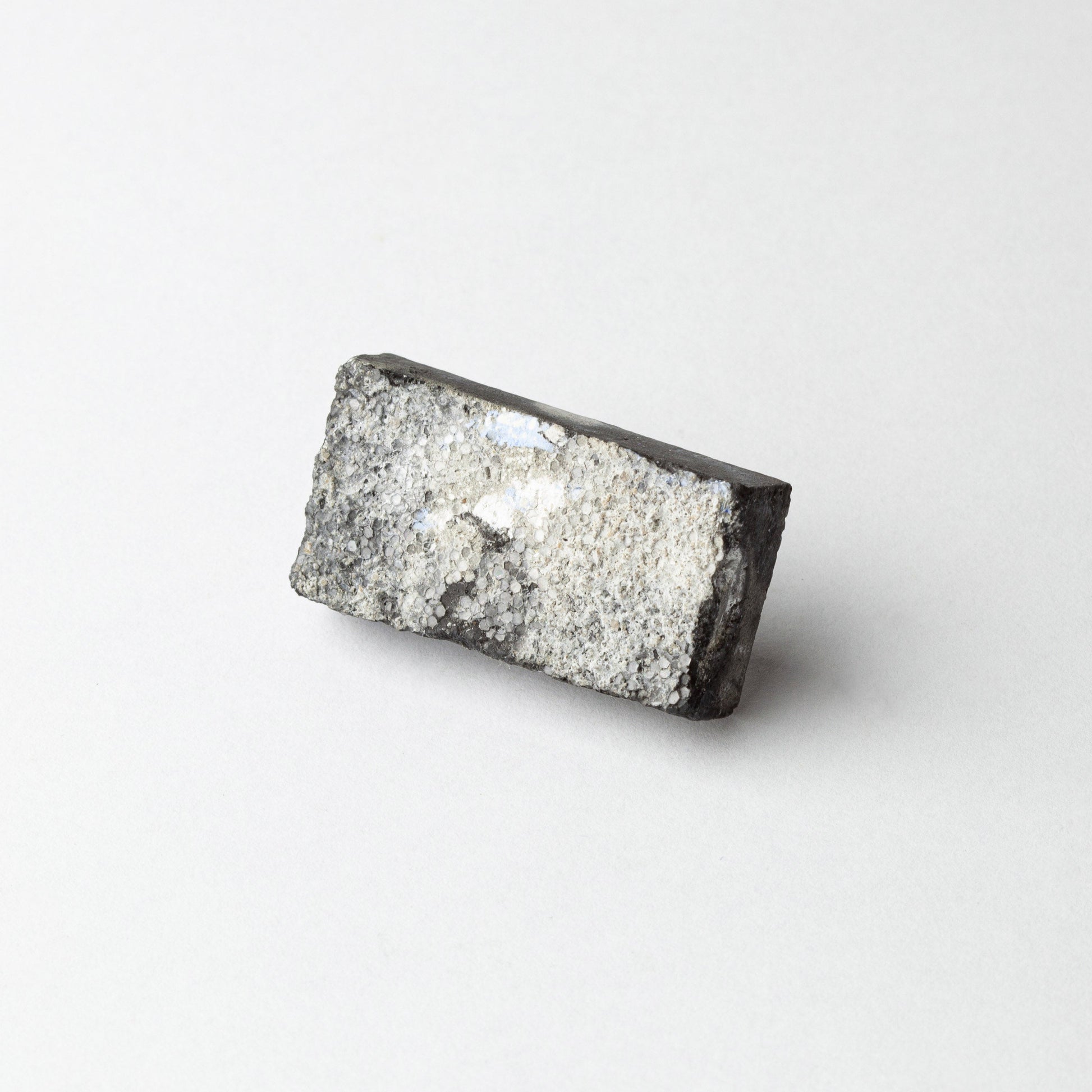 A rectangular gray and black textured stone or mineral sample with a rough, crystalline surface, photographed on a clean white background.