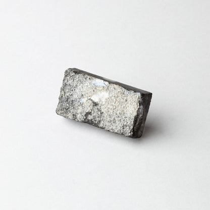 A rectangular gray and black textured stone or mineral sample with a rough, crystalline surface, photographed on a clean white background.