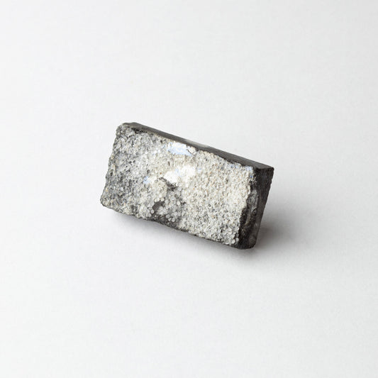 A rectangular gray and black textured stone or mineral sample with a rough, crystalline surface, photographed on a clean white background.