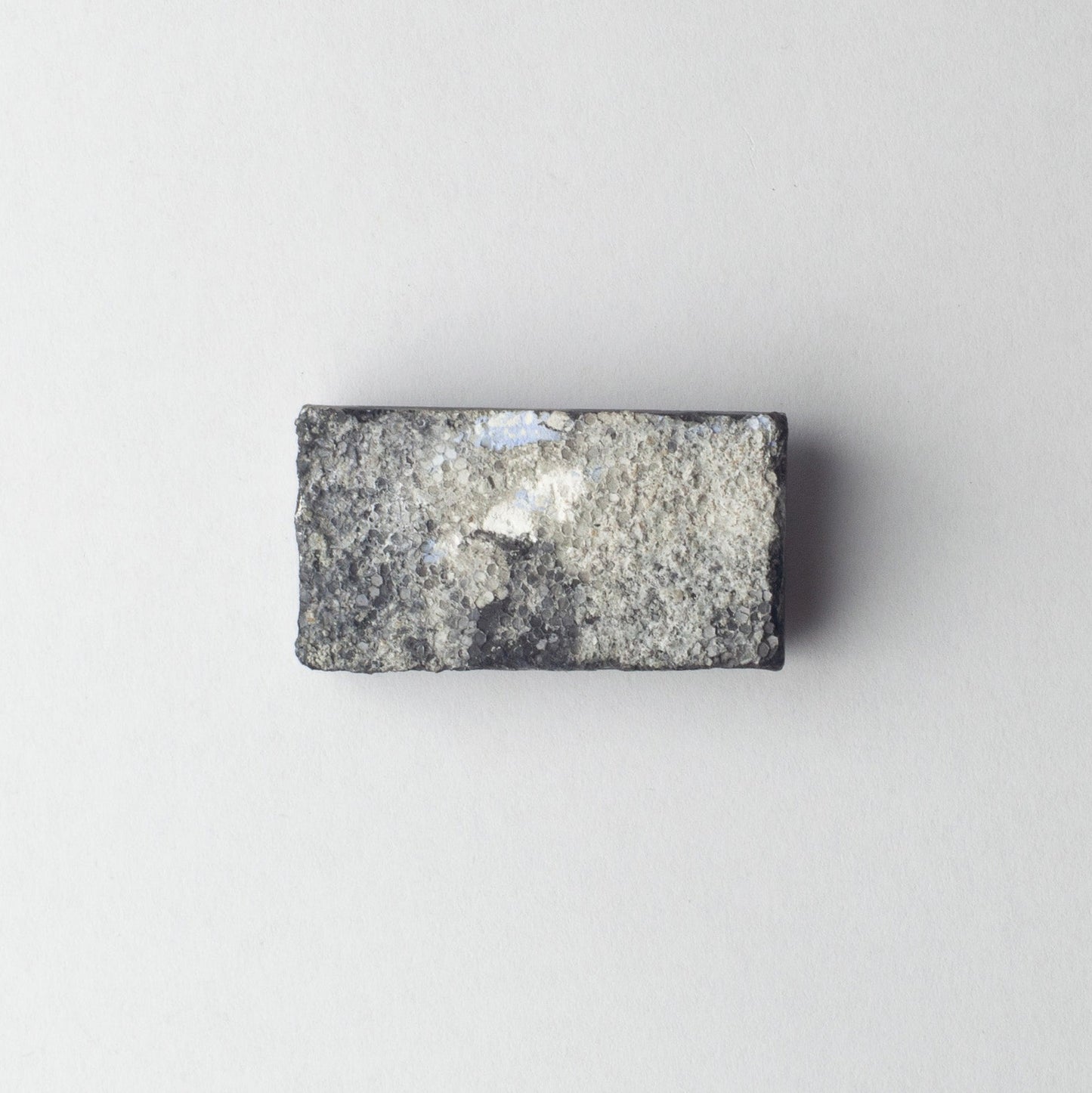 A rectangular gray stone or granite sample block with a rough, textured surface and a small white reflective patch, centered on a plain light gray background.