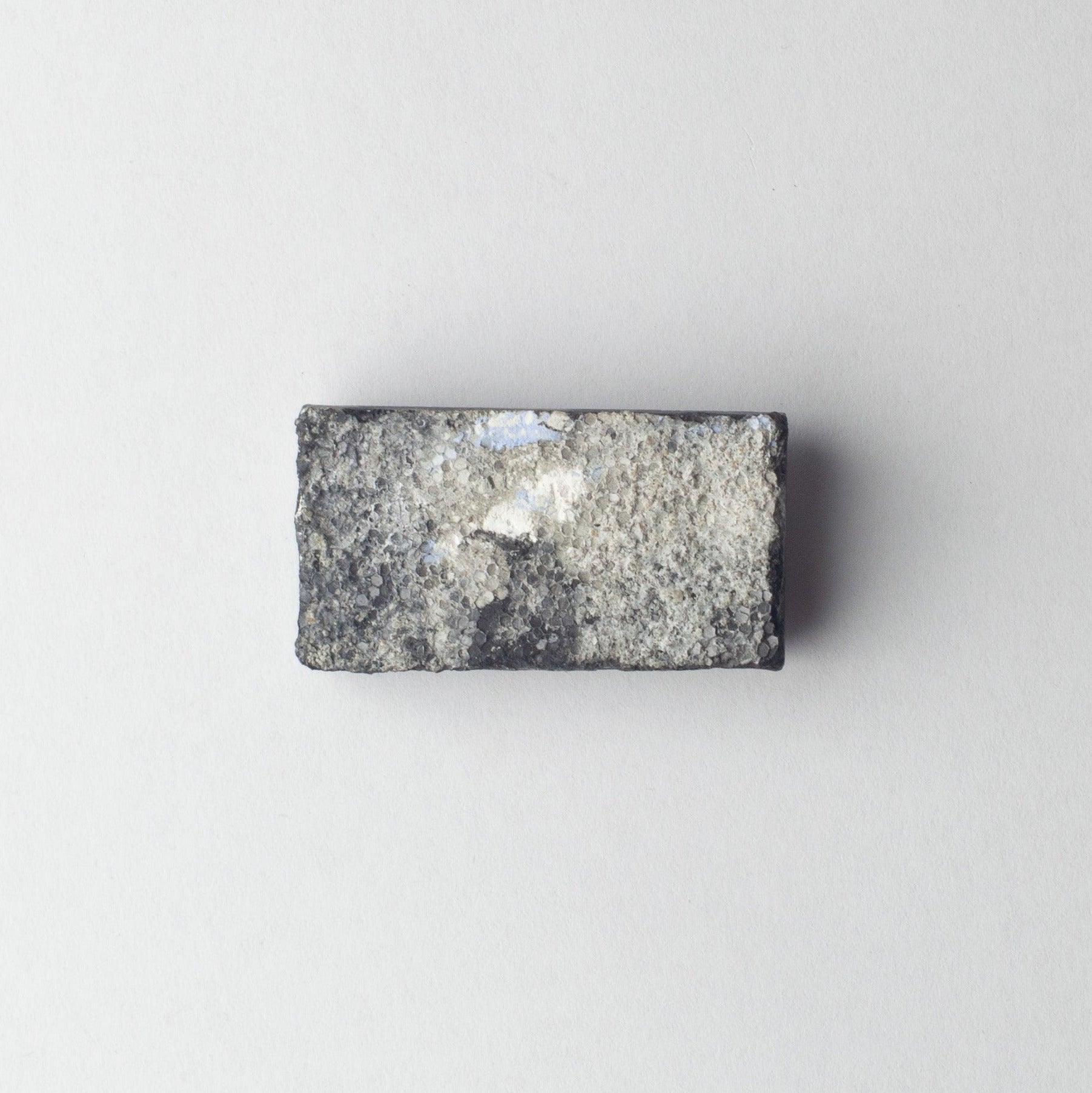 A rectangular gray stone or granite sample block with a rough, textured surface and a small white reflective patch, centered on a plain light gray background.