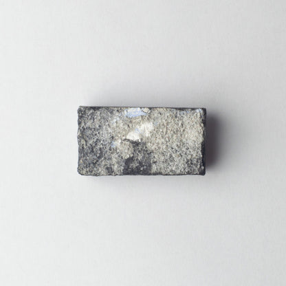 A rectangular gray stone or granite sample block with a rough, textured surface and a small white reflective patch, centered on a plain light gray background.