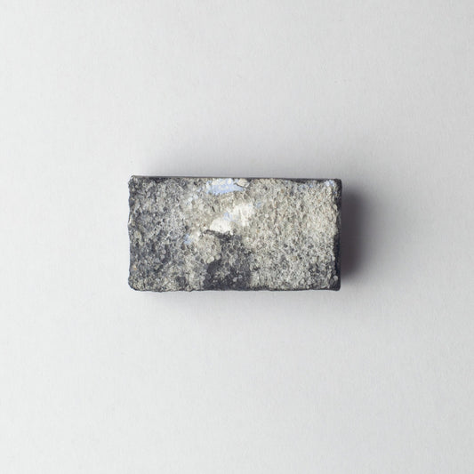 A rectangular gray stone or granite sample block with a rough, textured surface and a small white reflective patch, centered on a plain light gray background.