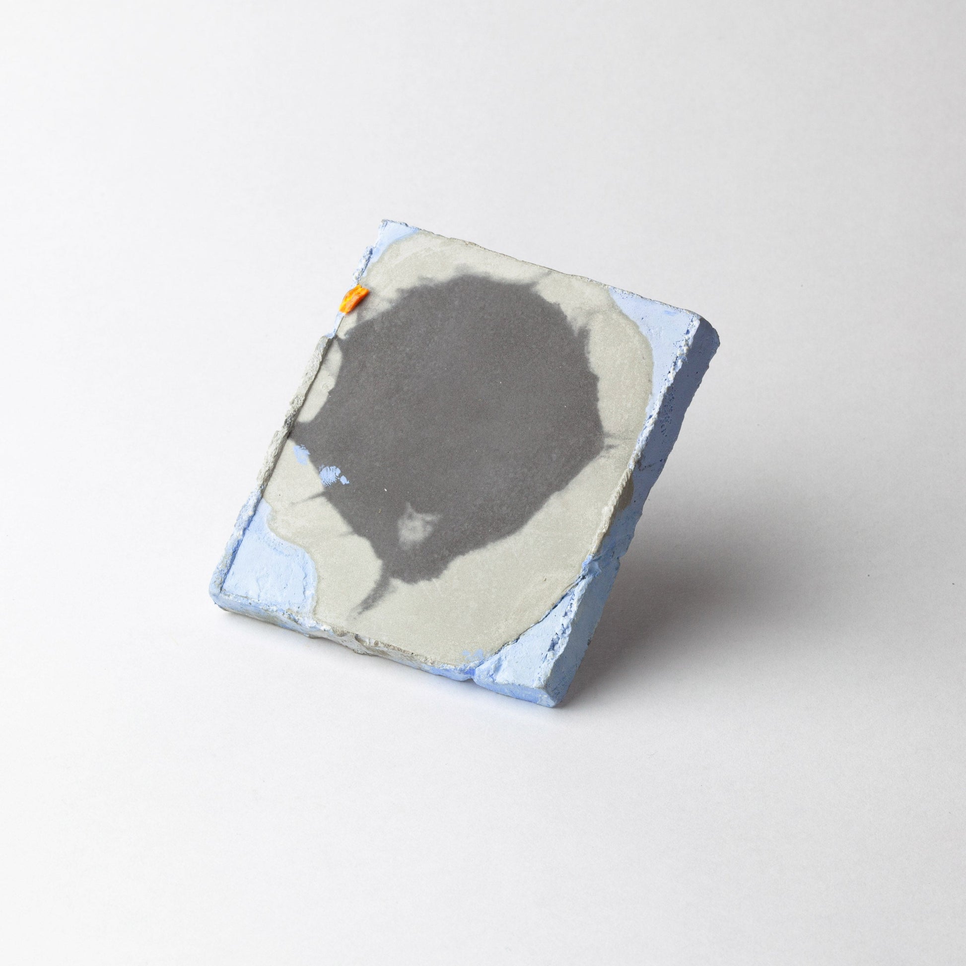 A square handmade ceramic brooch with a textured surface, featuring a dark gray abstract shape, light gray and white areas, light blue edges, and a small orange accent, set against a plain light gray background.