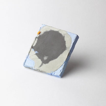 A square handmade ceramic brooch with a textured surface, featuring a dark gray abstract shape, light gray and white areas, light blue edges, and a small orange accent, set against a plain light gray background.
