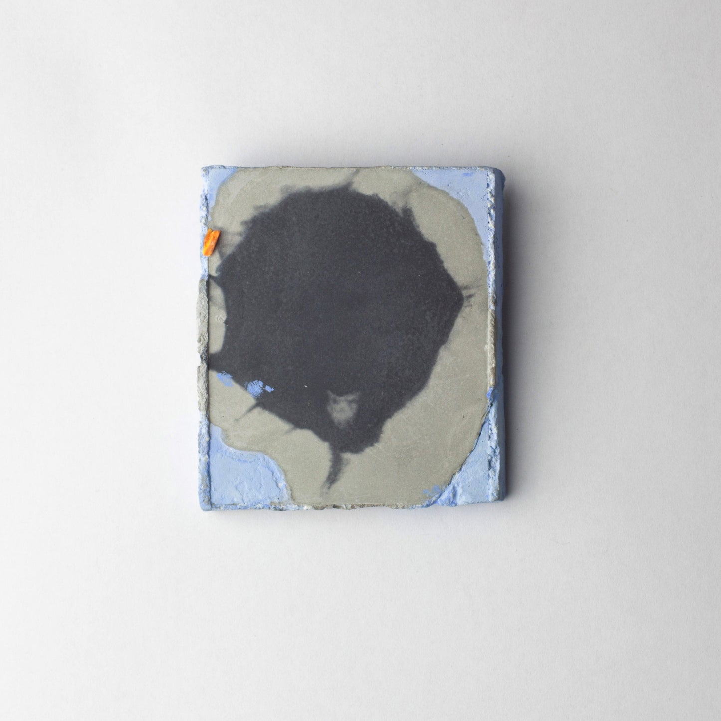 A square abstract brooch featuring a large black organic shape over a gray and light blue background, photographed against a clean light gray wall.