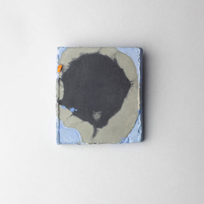 A square abstract brooch featuring a large black organic shape over a gray and light blue background, photographed against a clean light gray wall.