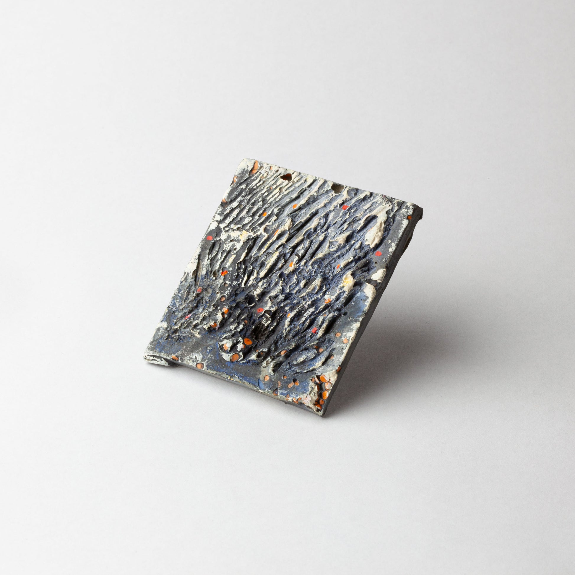 A square textured ceramic tile with a dark charcoal and gray surface, featuring organic raised patterns and small orange speckles, shown on a light gray background.