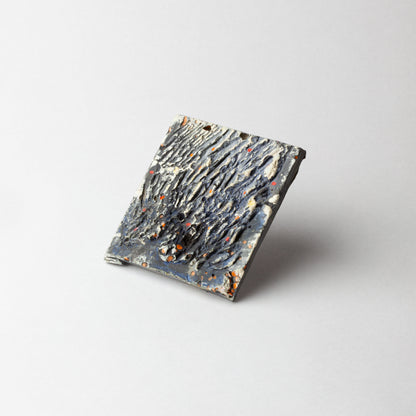 A square textured ceramic tile with a dark charcoal and gray surface, featuring organic raised patterns and small orange speckles, shown on a light gray background.