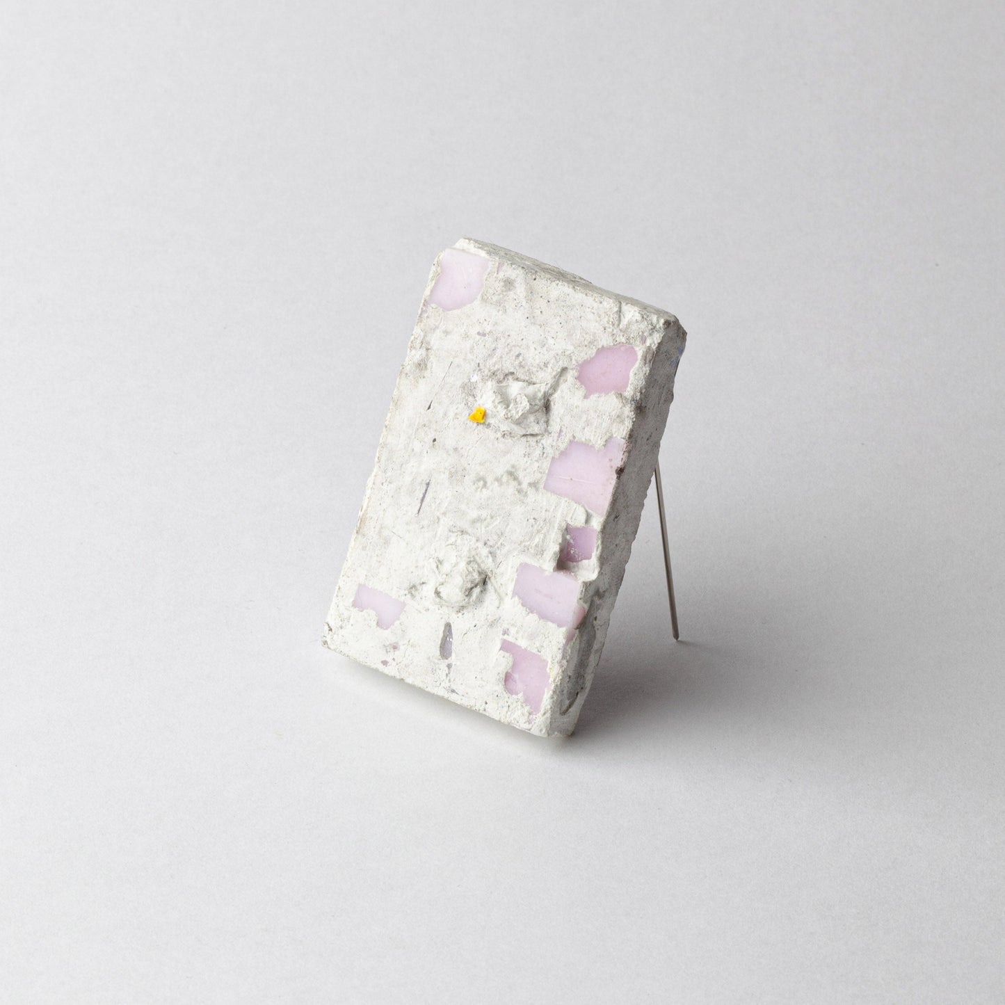 A rectangular concrete art piece with embedded light pink glass fragments and a small yellow fragment, displayed on a thin metal stand against a light gray background.