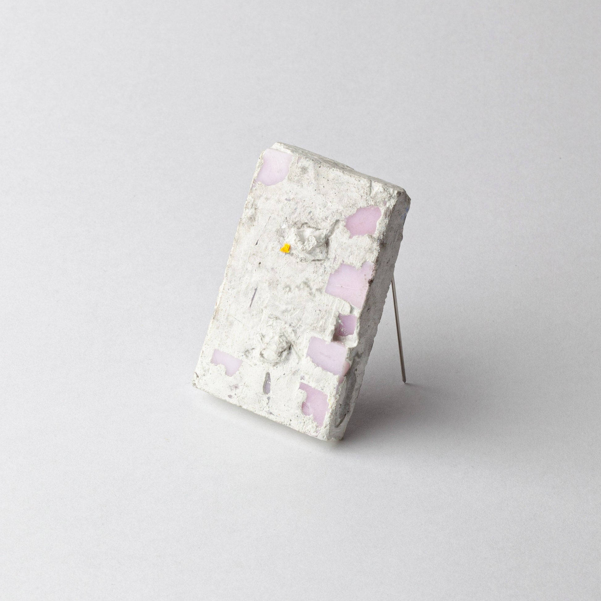 A rectangular concrete art piece with embedded light pink glass fragments and a small yellow fragment, displayed on a thin metal stand against a light gray background.