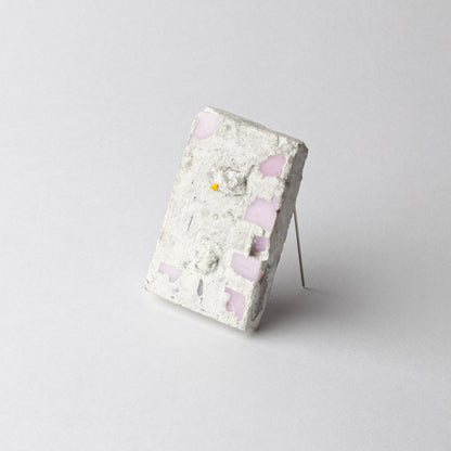 A rectangular concrete art piece with embedded light pink glass fragments and a small yellow fragment, displayed on a thin metal stand against a light gray background.