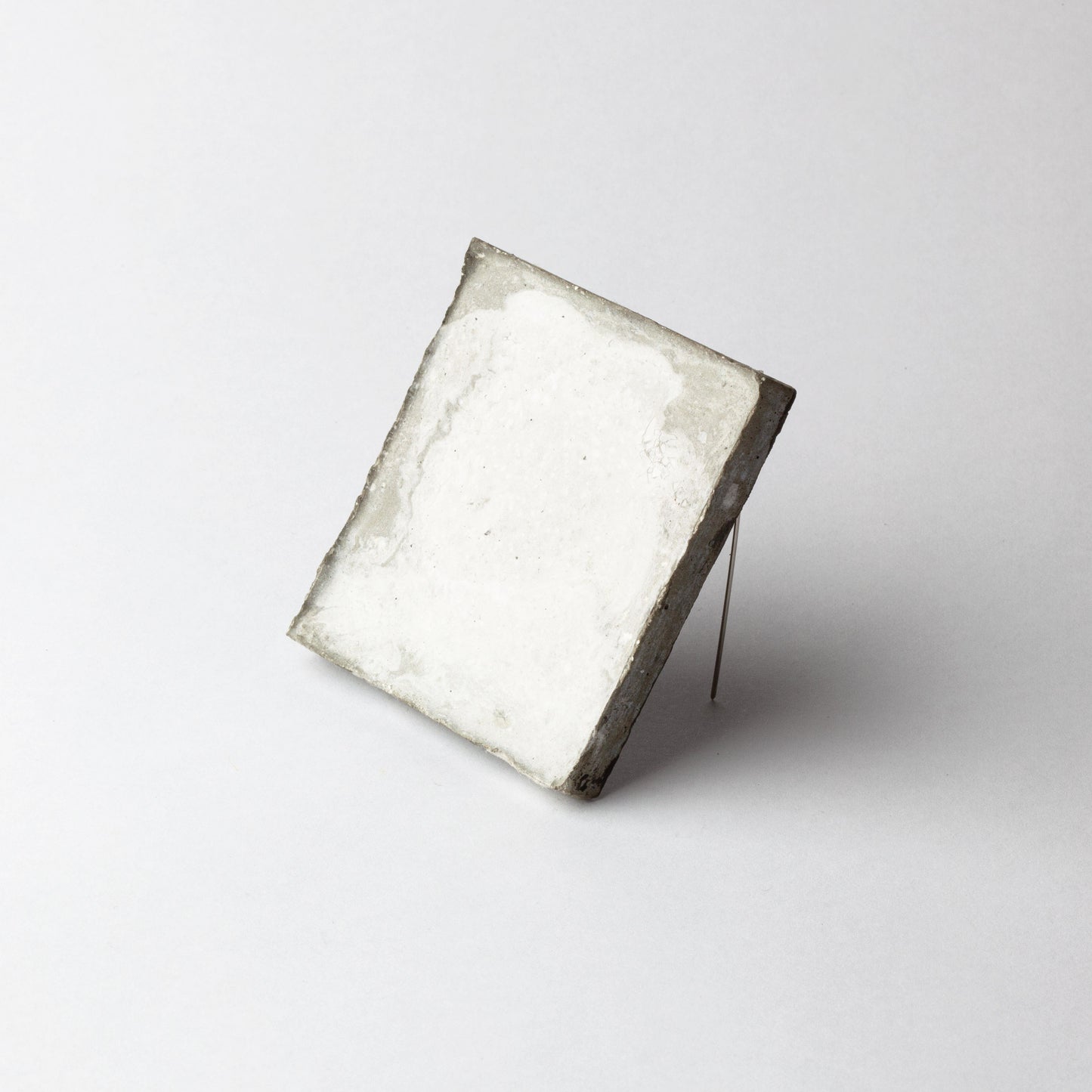 A square white textured tile or coaster with rough, rustic gray edges, propped up on a thin metal wire stand against a clean white background.