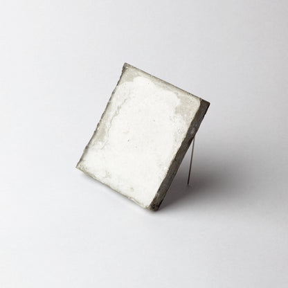 A square white textured tile or coaster with rough, rustic gray edges, propped up on a thin metal wire stand against a clean white background.