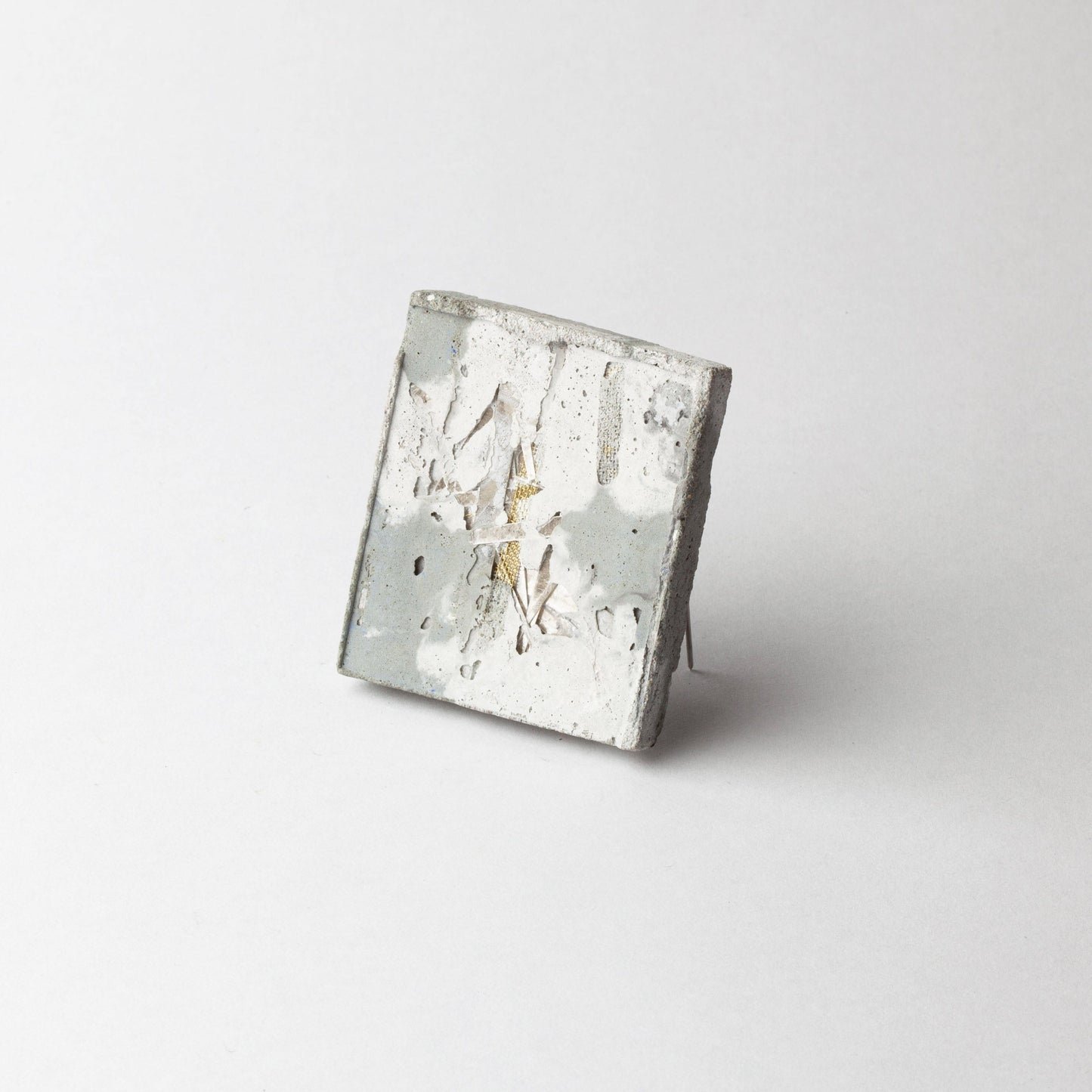 A square concrete art piece with a distressed, weathered finish in shades of gray, white, and subtle gold-toned highlights, photographed on a seamless light gray background.