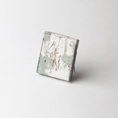 A square concrete art piece with a distressed, weathered finish in shades of gray, white, and subtle gold-toned highlights, photographed on a seamless light gray background.